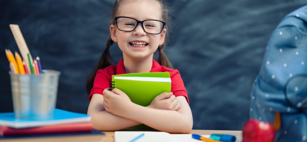 Back to school! Happy cute industrious child is sitting at a desk indoors. Kid is learning in class on background of blackboard.