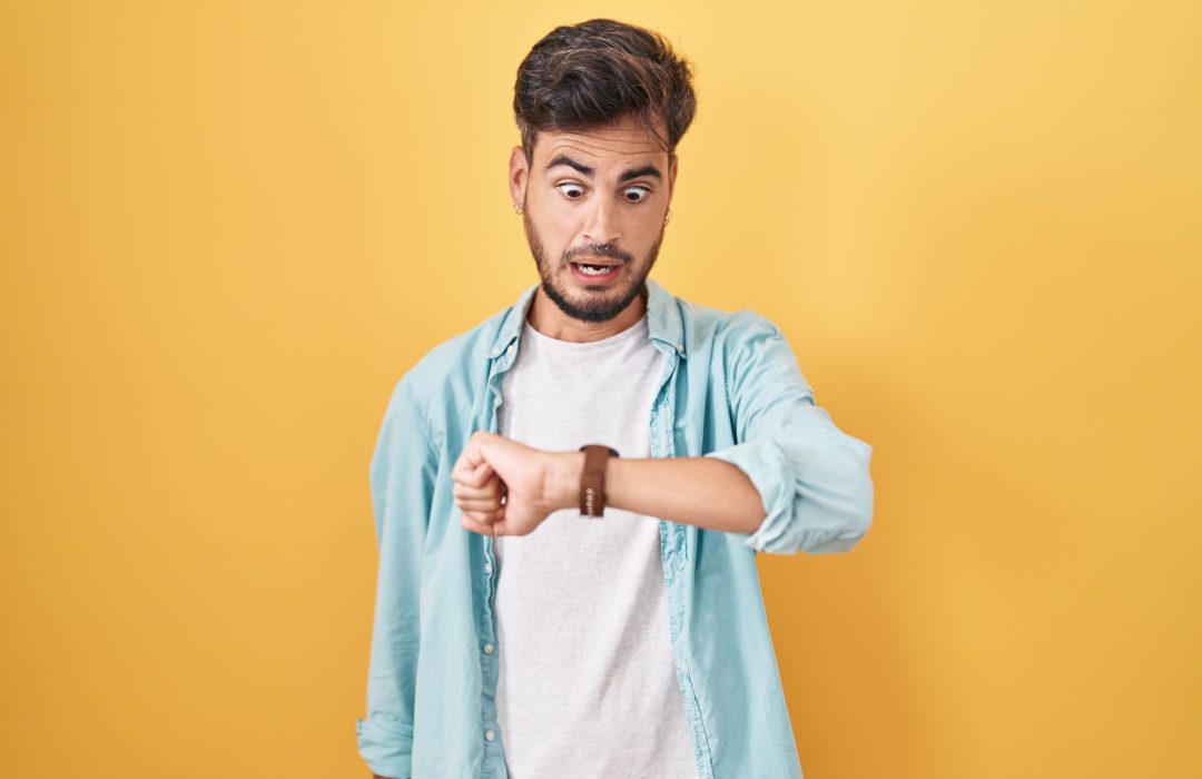 Young hispanic man with tattoos standing over yellow background looking at the watch time worried, afraid of getting late